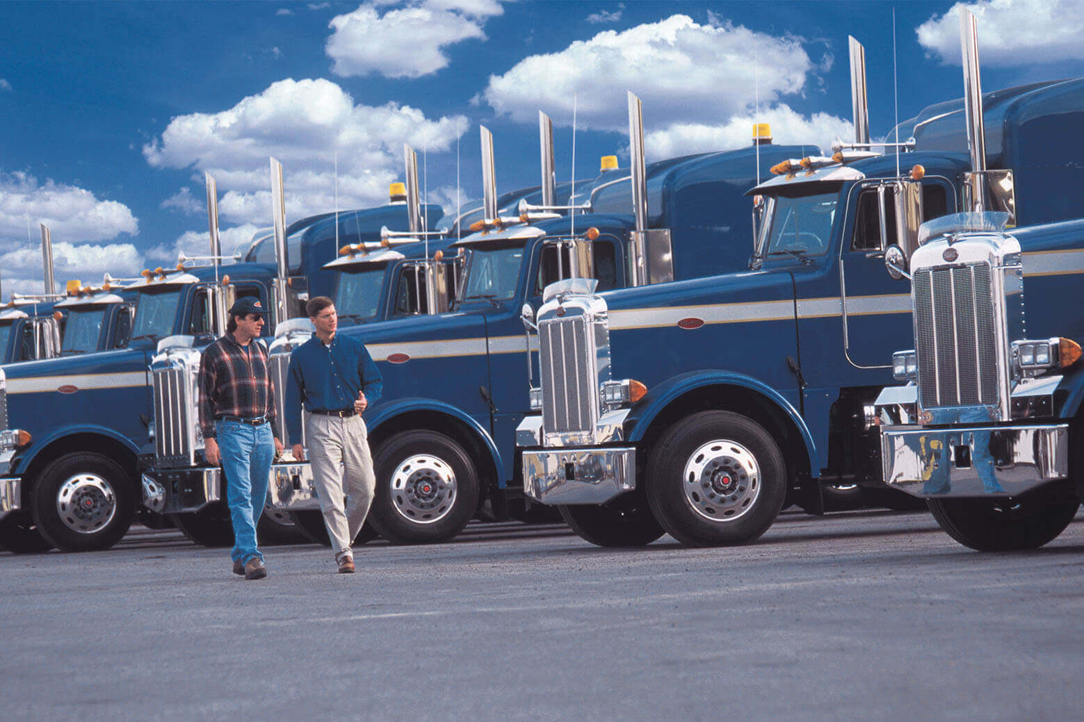 Two men walking in front of a fleet of blue semi trucks.