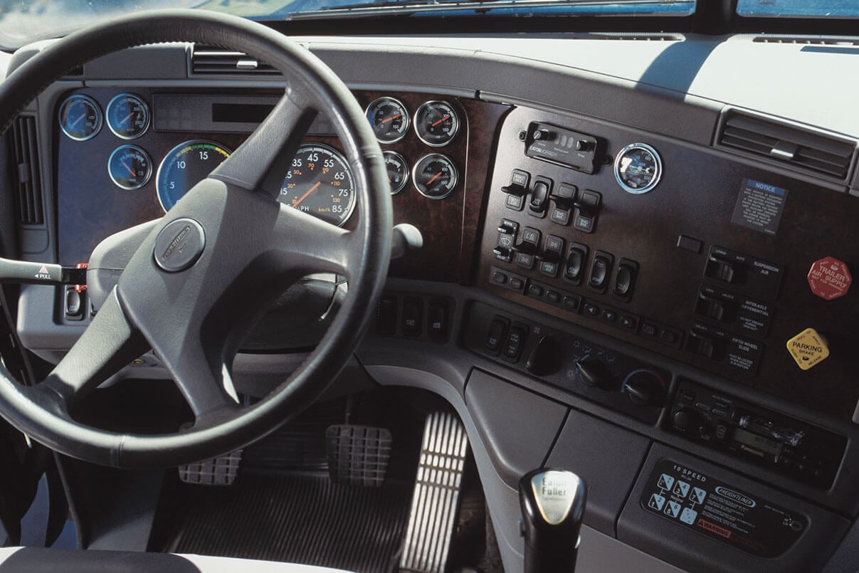 The interior dash of a semi truck showing the steering wheel, gauges, and switches.